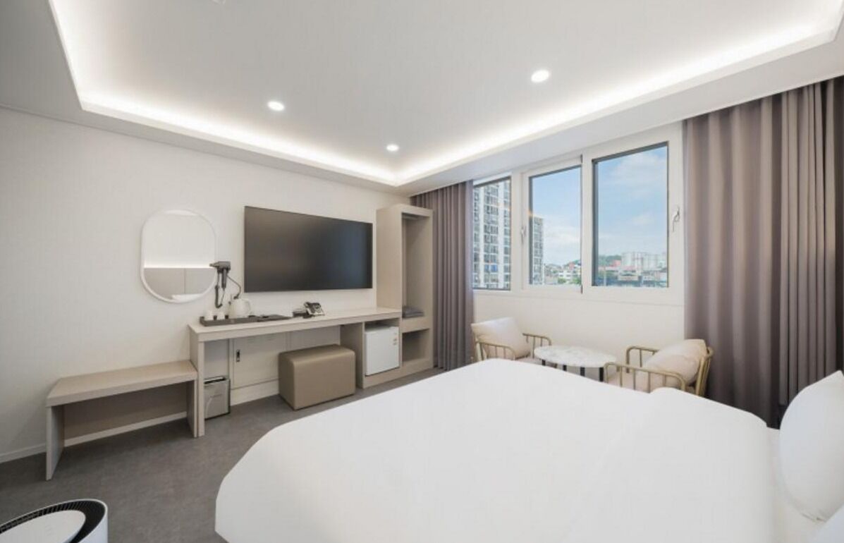 A hotel room with a bed, desk, chair, TV, and large windows.
