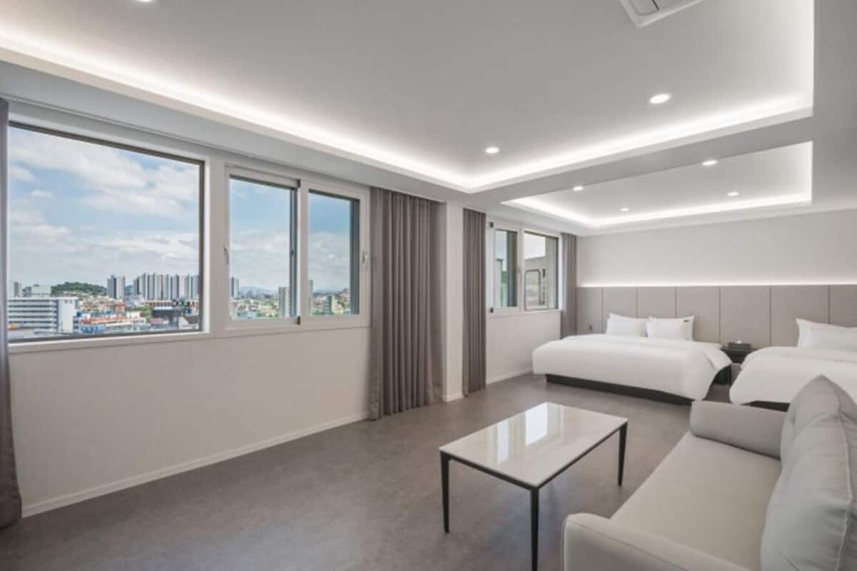 Una habitación de hotel moderna con una cama grande, un sofá, una mesita de centro y vistas a la ciudad a través de amplias ventanas.