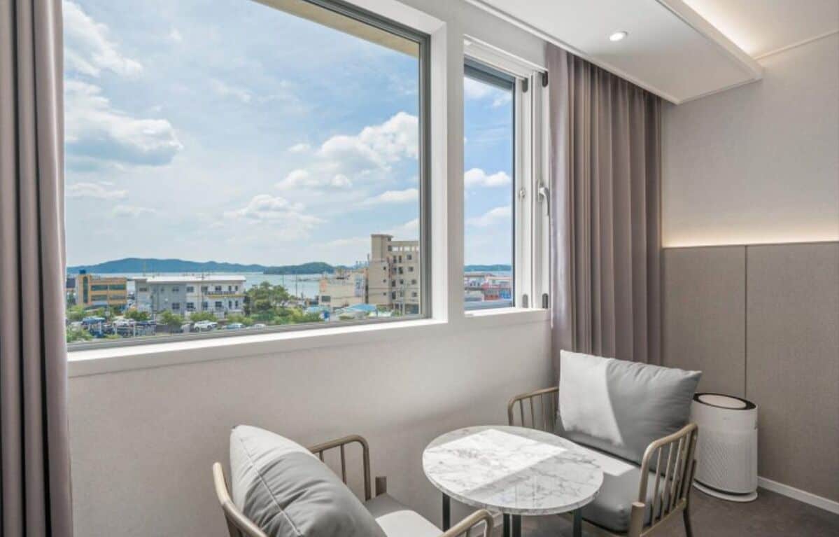 A hotel room with a large window, a round table, a sofa, and a trash bin.
