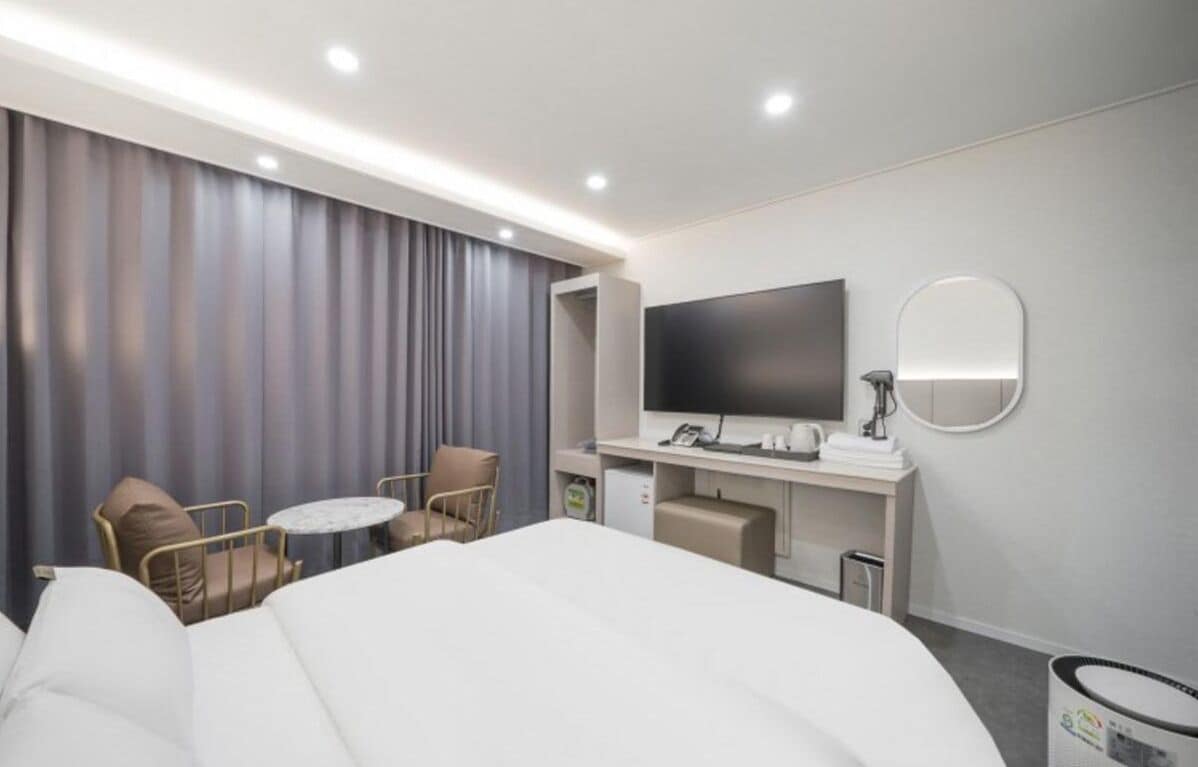 A hotel room with a bed, a desk with a flat-screen TV, a round mirror, and two chairs.