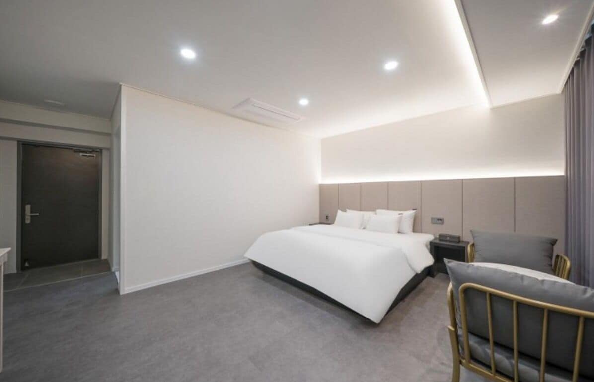 A modern hotel room with a large bed, bedside tables, a chair, and a door.