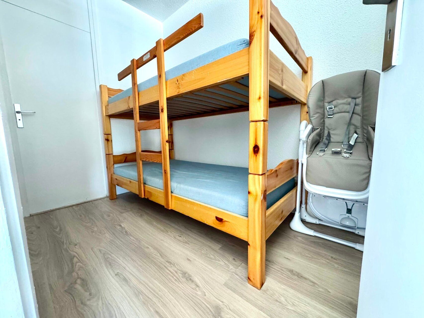 A bunk bed with a blue mattress, a wooden frame, and a wheelchair to the right.