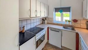 Cocina compacta con armarios blancos, encimera negra, estufa, lavavajillas y ventana con vistas al verdor.