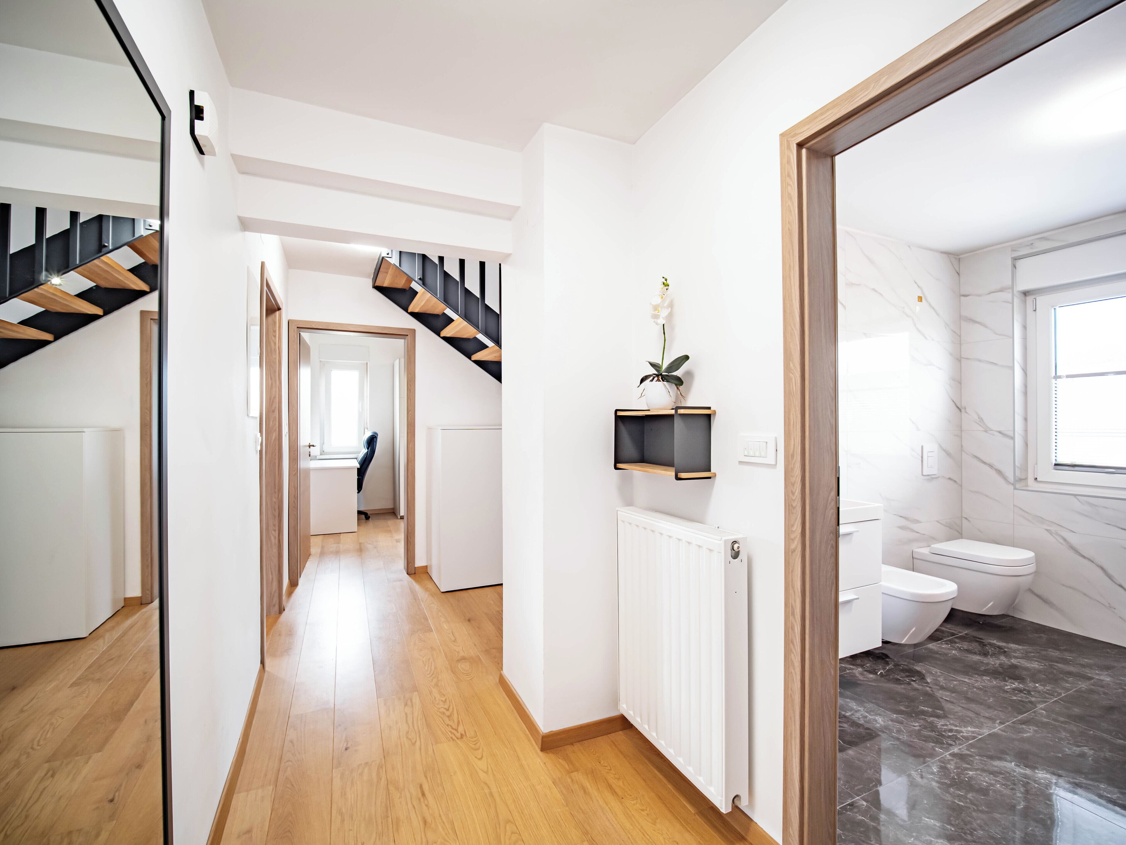 A modern interior with a wooden staircase, a hallway leading to a room, a radiator, and a bathroom with a toilet and marble flooring.