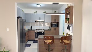 A modern kitchen with a built-in oven, microwave, and a gas stove. There are two wooden bar stools and a small dining table. - Boho Luxury Stay•Pool•Gym/Near Everything (Santiago de los Caballeros)
