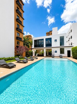 A swimming pool with lounge chairs, a modern building with balconies, and a clear blue sky. - Boho Luxury Stay•Pool•Gym/Near Everything (Santiago de los Caballeros)