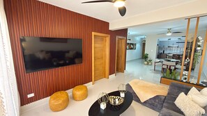 A modern living room with a flat-screen TV, wooden panel walls, a sofa, ottomans, and a coffee table with decorative items. - Boho Luxury Stay•Pool•Gym/Near Everything (Santiago de los Caballeros)