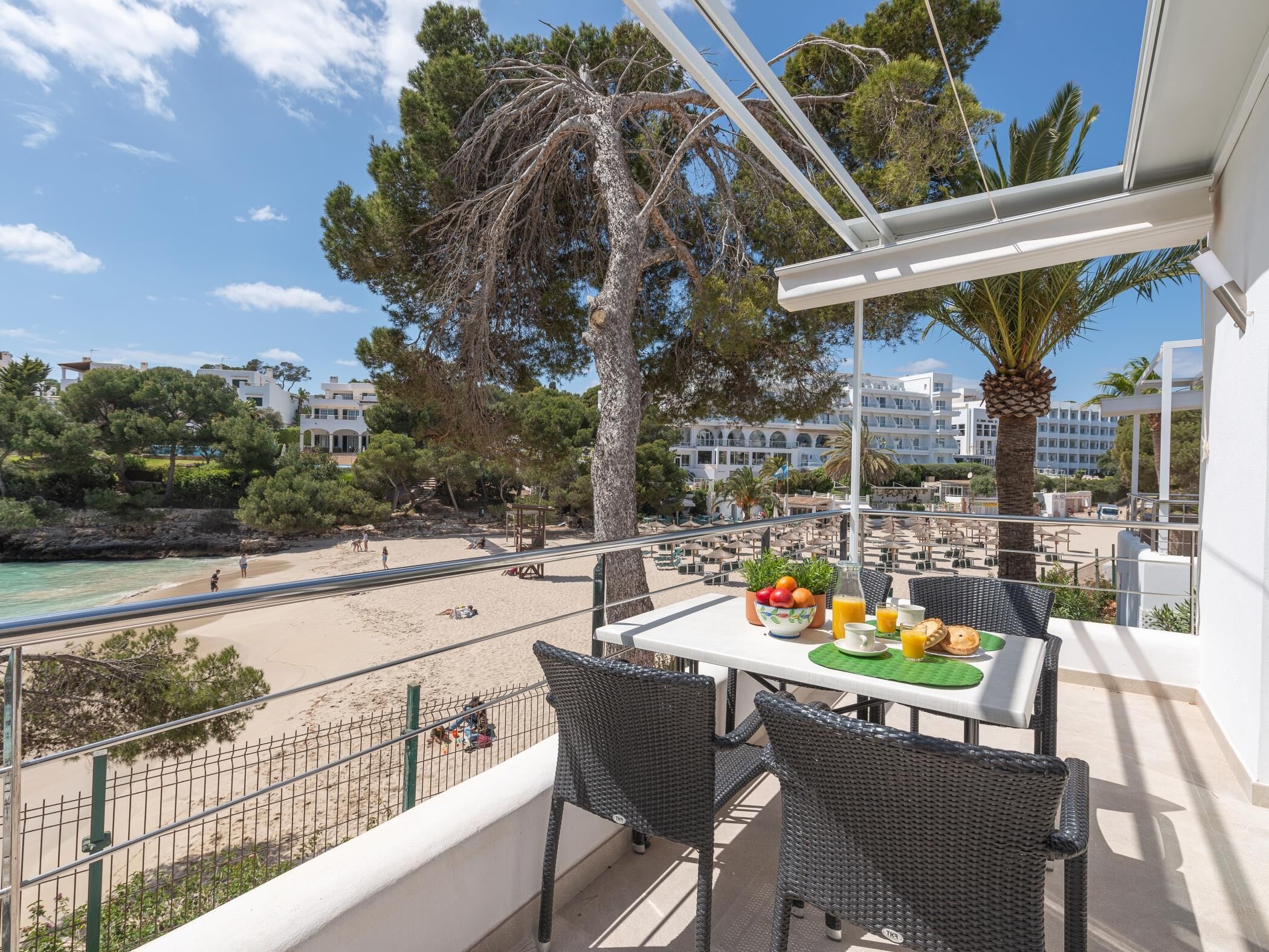 A balcony with wicker chairs, a table with fruit and drinks, overlooking a beach and a resort area.