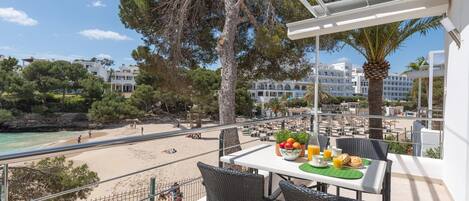 A balcony with wicker chairs, a table with fruit and drinks, overlooking a beach and a resort area.
