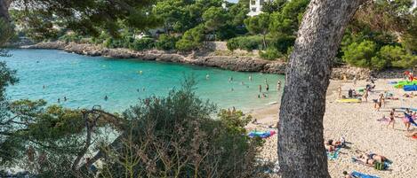 Una playa con agua turquesa cristalina, gente nadando y tomando el sol, y edificios al fondo.