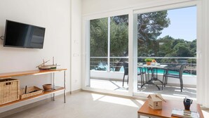 A modern room with a TV, a wooden table, a wicker shelf, and a view of a pool and trees. - Apartment Playa Dor 27 in first sea line, Cala Gran beach (Cala D'or (Santanyí))