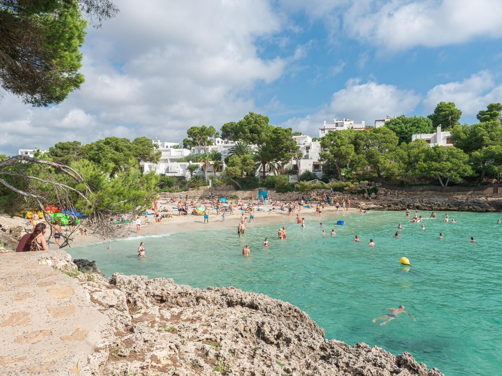Uma praia com águas turquesa cristalinas, costa rochosa e edifícios brancos ao fundo.