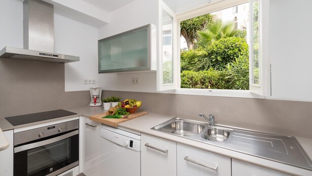 A modern kitchen with white cabinets, stainless steel appliances, a built-in oven, a dishwasher, and a window with a view of greenery. - Apartment Playa Dor 27 in first sea line, Cala Gran beach (Cala D'or (Santanyí))