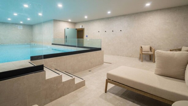 Une piscine intérieure moderne avec des parois en verre transparent, un espace détente avec un canapé et des fauteuils, et un sol carrelé.