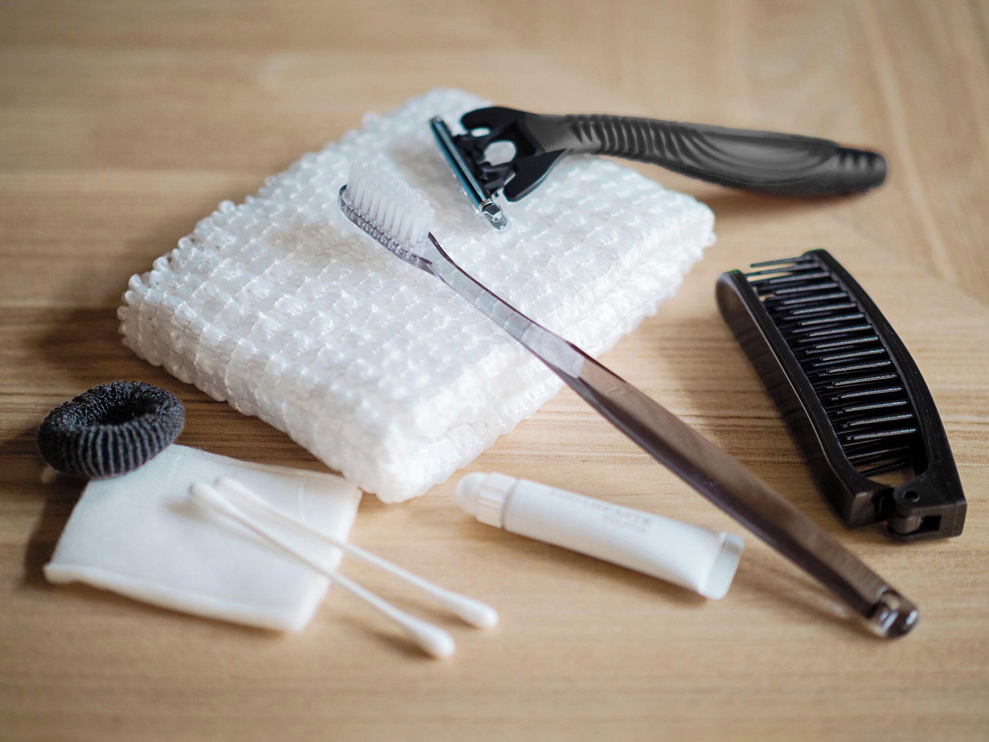 A wooden surface with a white textured object, a black comb, a metal brush, a black cylindrical object, a white tube, and white cotton swabs.