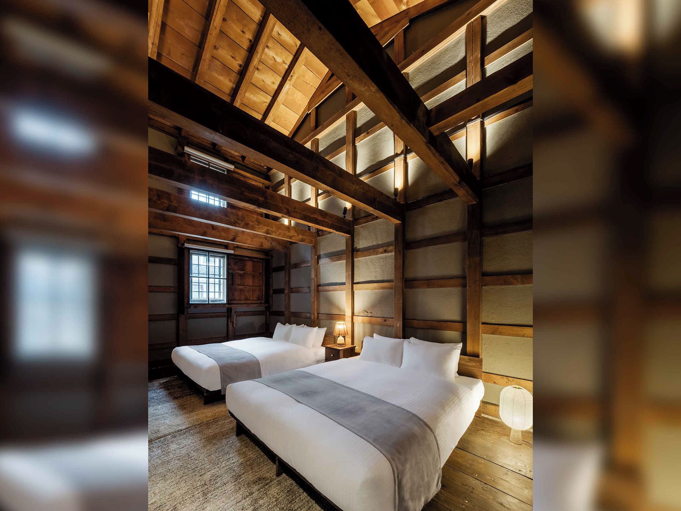 Una habitación con vigas de madera, una cama grande, una cama más pequeña, un ventanal y una lámpara.