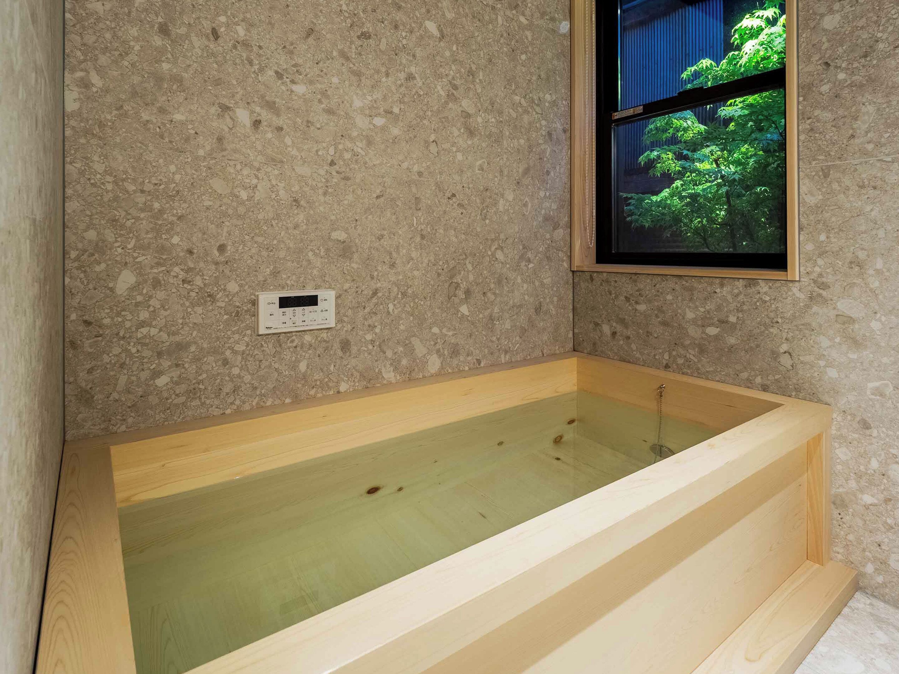 A wooden bathtub in a room with a speckled wall and a window with a view of greenery.