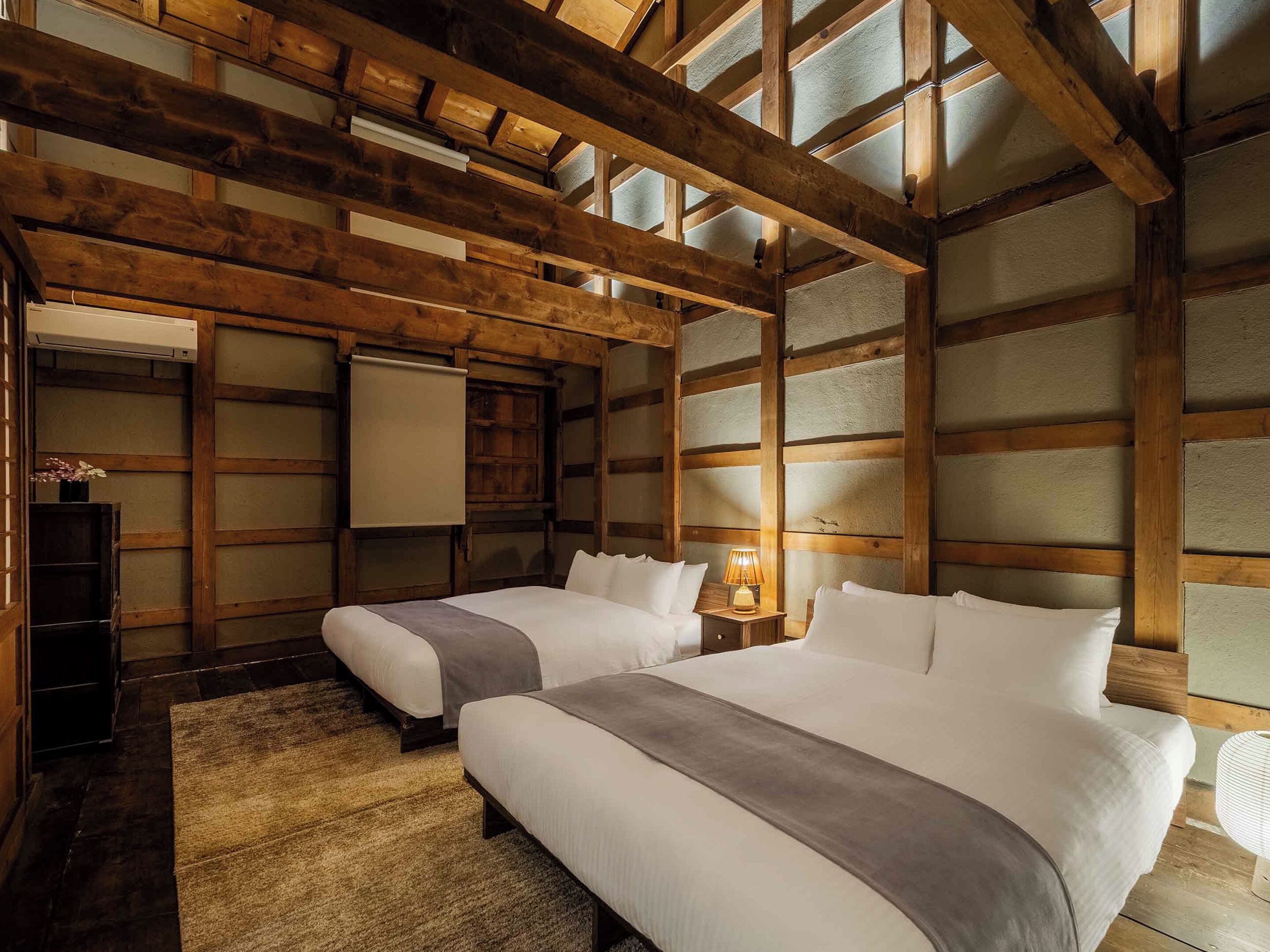Um quarto com vigas de madeira, duas camas com roupa de cama branca, uma mesa pequena com abajur e um ar condicionado fixado na parede.