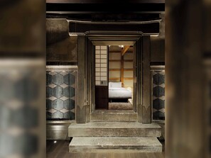 Ein traditioneller japanischer Raum mit Tatami-Bodenbelag, einem niedrigen Holzbett und einer verschiebbaren Tür zu einem weiteren Zimmer.