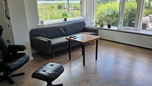 Une pièce spacieuse avec un canapé gris, une table basse en bois, un fauteuil en cuir noir et un pouf noir. De grandes fenêtres offrent une vue sur la verdure et un plan d’eau.
