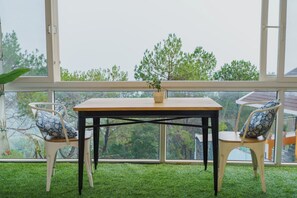Una habitación con una mesa, dos sillas, una planta en maceta y vistas a árboles a través de ventanas amplias.