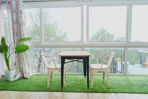 A room with a large window, a wooden table, two chairs, and a potted plant. - Acanti Dharamshala (Dharamshala)