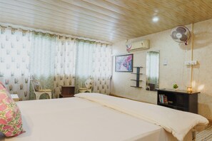 Un dormitorio con una cama, una silla, un escritorio, un ventilador y una estantería con libros y una planta.