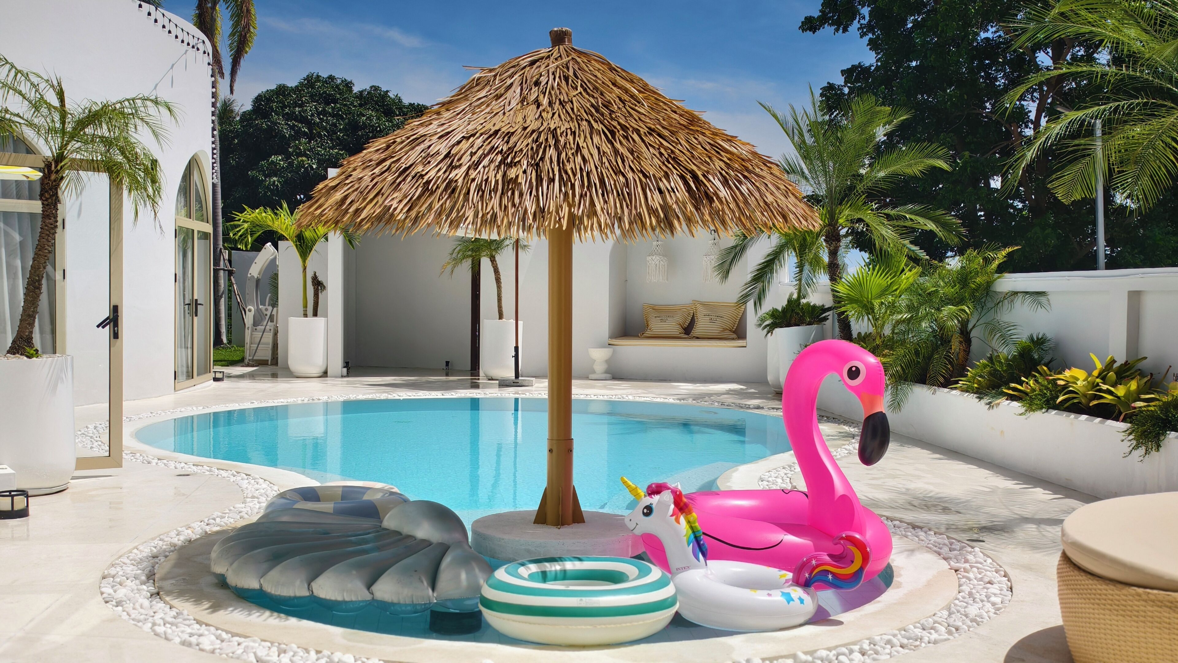 A swimming pool with a thatched umbrella, inflatable rings, and a unicorn float.