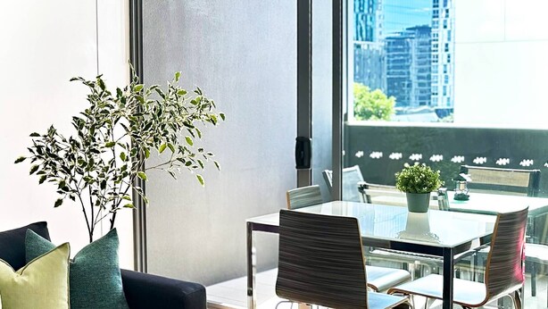 Un soggiorno moderno con divano, tavolo da pranzo con sedie, una pianta in vaso e un'ampia finestra con vista sulla città.