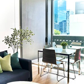 A modern living room with a sofa, a dining table with chairs, a potted plant, and a large window with a city view. - MAANGO - 1BR retreat• James St • Free Parking • Balcony • Pool (Fortitude Valley)
