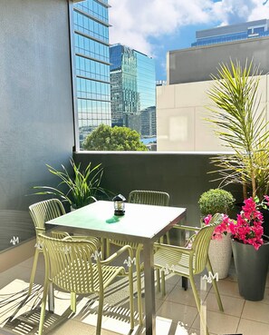 Ein kleiner Sitzbereich im Freien mit Tisch und Stühlen, einem Blumentopf und Blick auf eine Stadt durch ein Fenster.