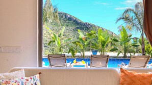 A room with a sofa, a view of a pool with chairs, and potted plants. - Villa Fonte, outstanding view, large pool (İslamlar)