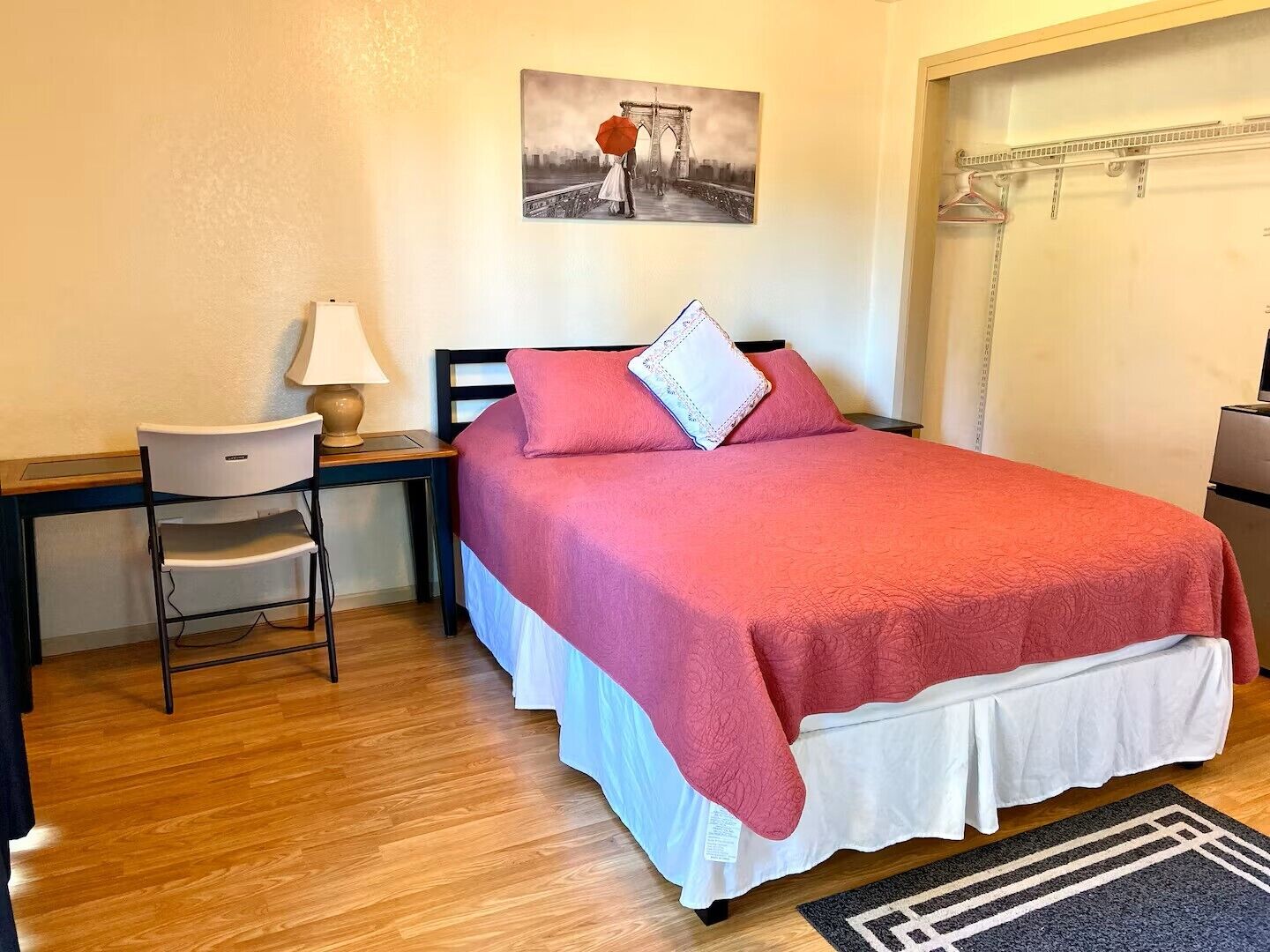 A hotel room with a bed, a desk, a chair, a lamp, a painting, and a shower area.