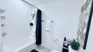 Un bagno moderno con doccia rivestita di piastrelle bianche, una vasca da bagno, un WC e un lavandino. Sul pavimento ci sono tappetini neri, un asciugamano appeso al muro e una pianta sul lavandino.