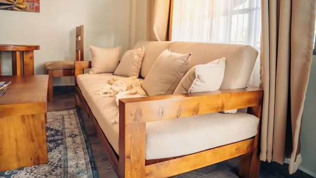 A wooden sofa with cushions, a wooden side table, and a patterned rug in a well-lit room with curtains. - Lights & Kulture Homes a 2-bedroom apartment for creatives in Arusha Region (Arusha Region)