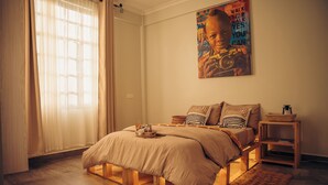 A bedroom with a bed, a window with curtains, a wall-mounted poster, a bedside table, and a nightstand. - Lights & Kulture Homes a 2-bedroom apartment for creatives in Arusha Region (Arusha Region)