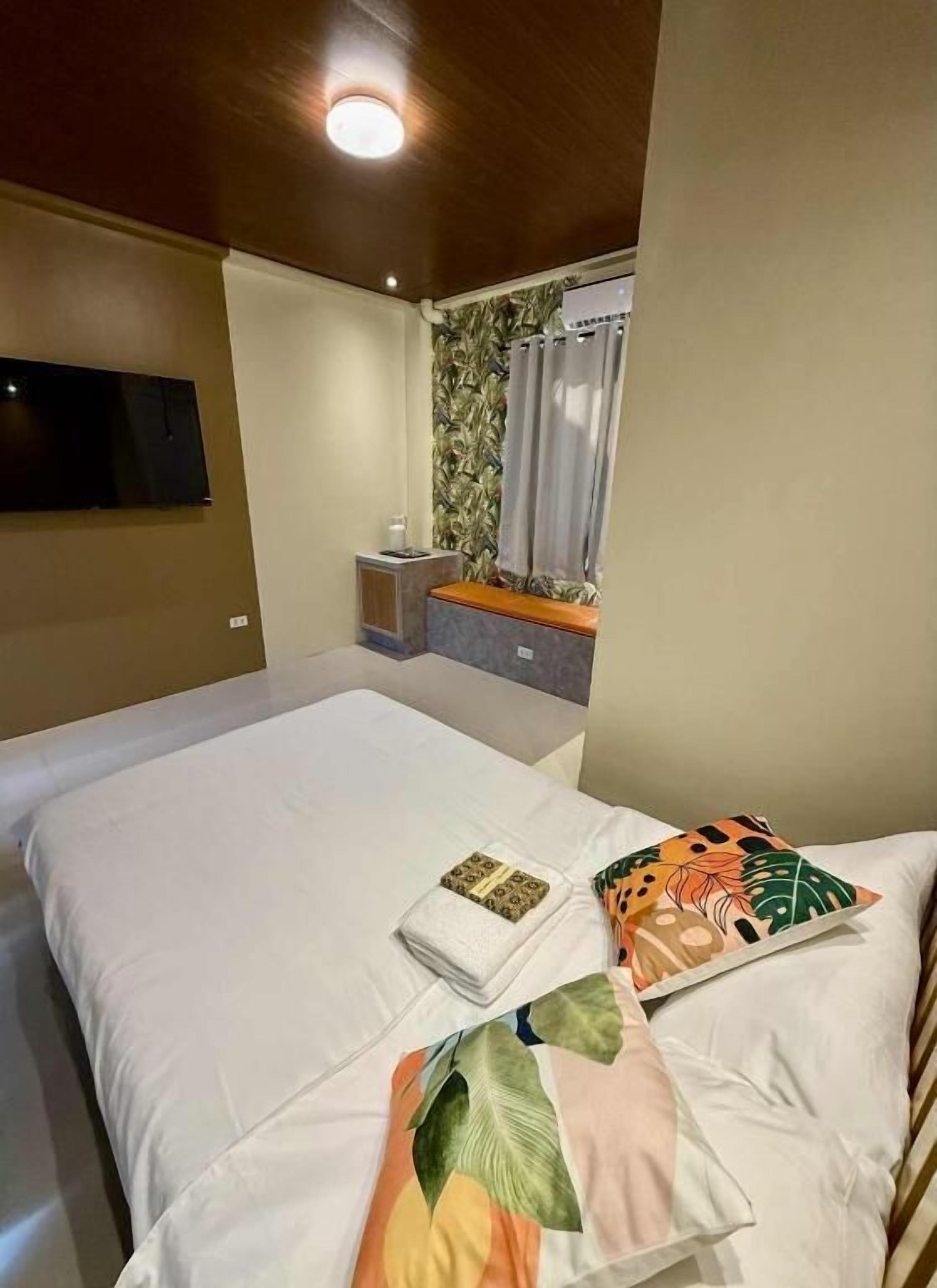 A hotel room with a bed, a TV mounted on the wall, a small bedside table, and a window with curtains.