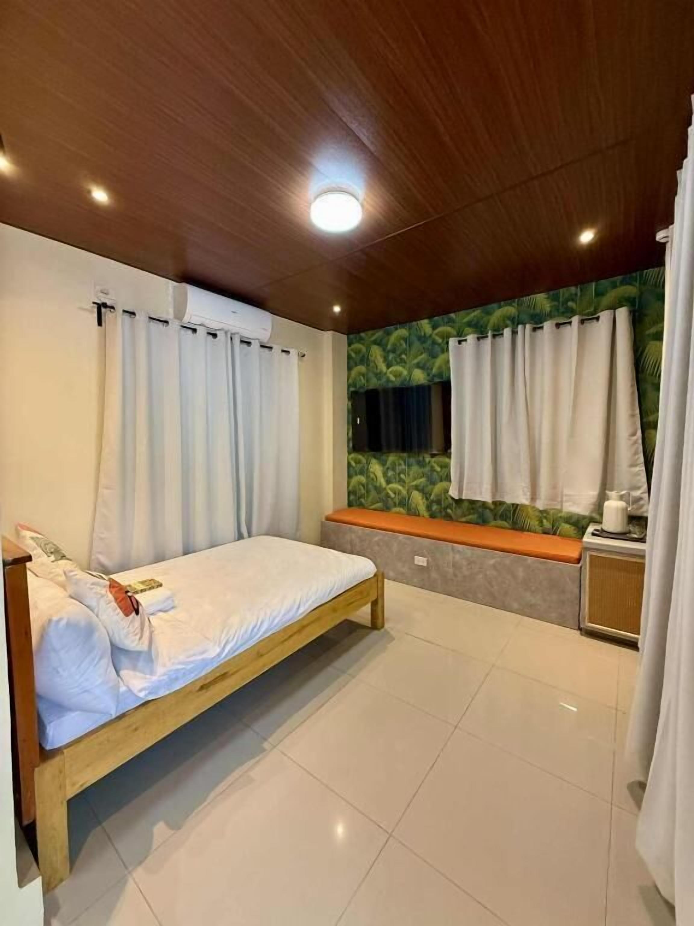 Un dormitorio con cama de madera, cortinas blancas, una ventana con vistas y suelo de baldosas.