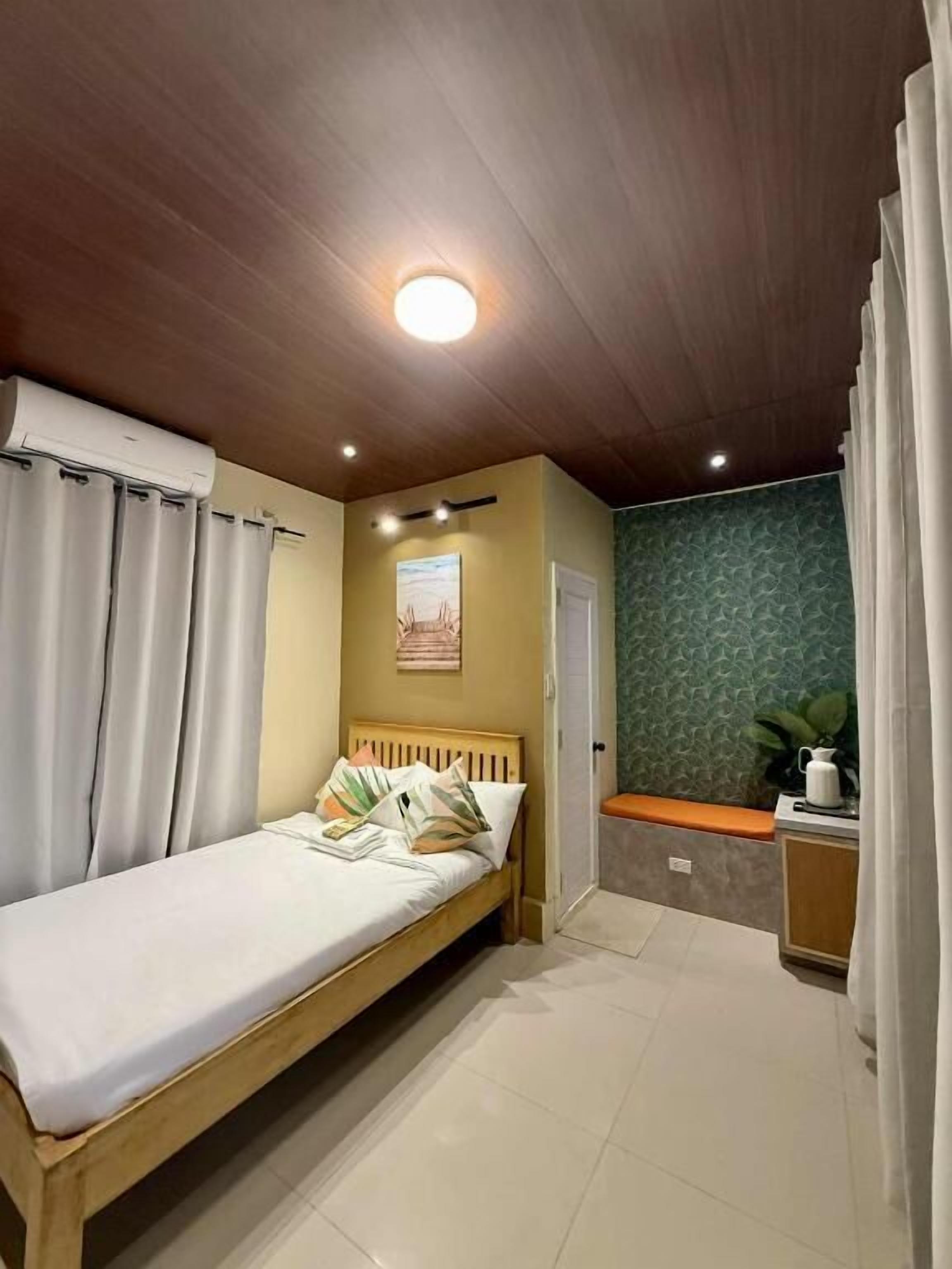 A bedroom with a wooden bed, a small bedside table, a green patterned wall, and a ceiling light.