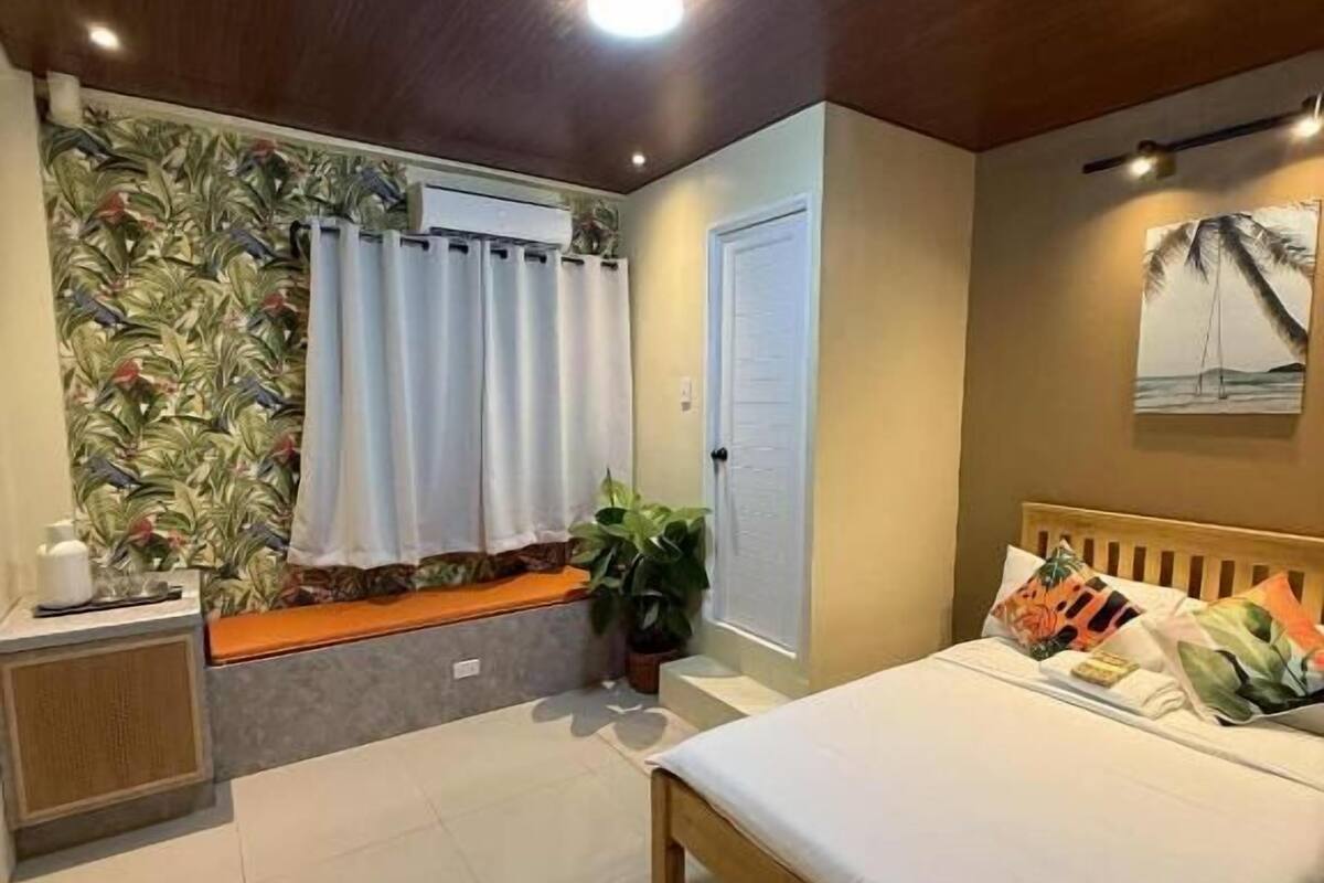 A bedroom with a bed, a window with curtains, a plant, and a door.