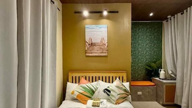 A hotel room with a bed, curtains, a headboard, a small table with a plant, and a painting on the wall.