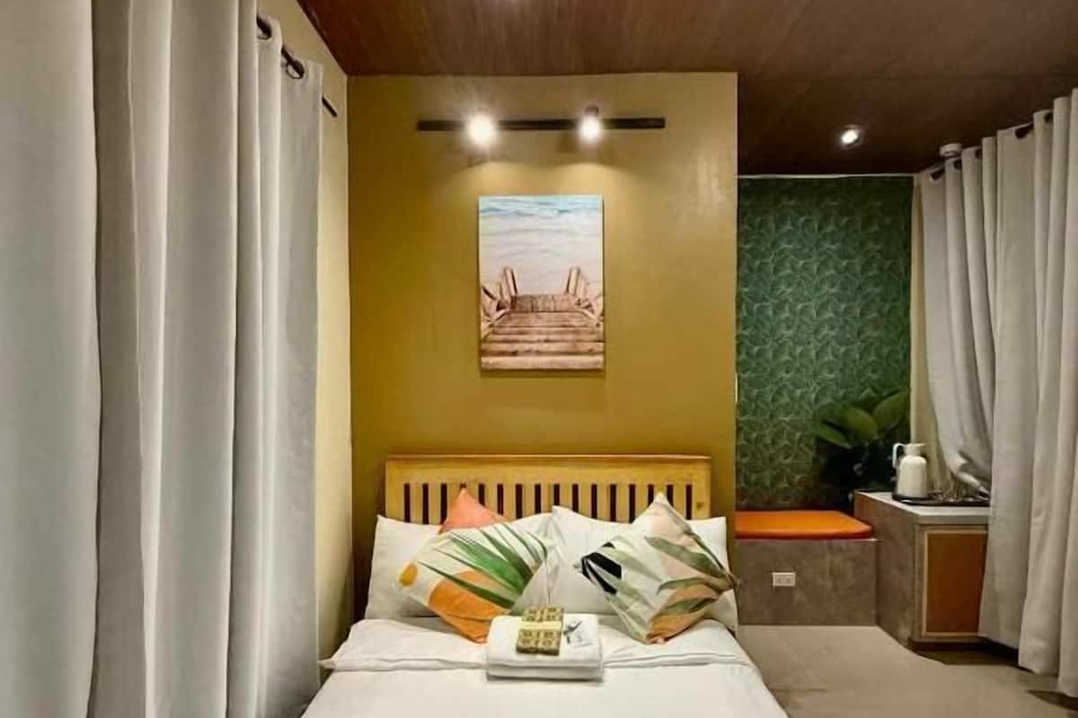 A hotel room with a bed, curtains, a headboard, a small table with a plant, and a painting on the wall.