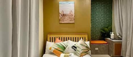 A hotel room with a bed, curtains, a headboard, a small table with a plant, and a painting on the wall.