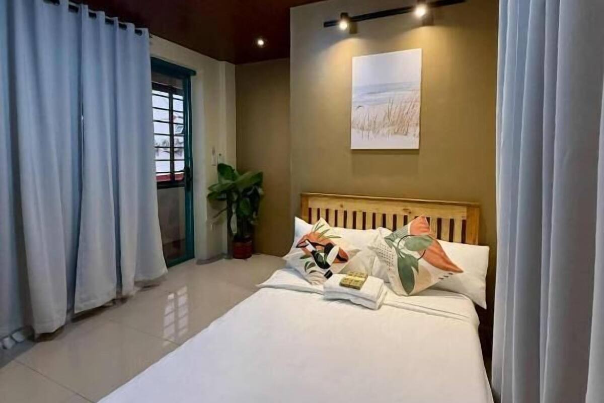 A bedroom with a wooden headboard, a bed with white bedding, a wall-mounted artwork, and a potted plant.