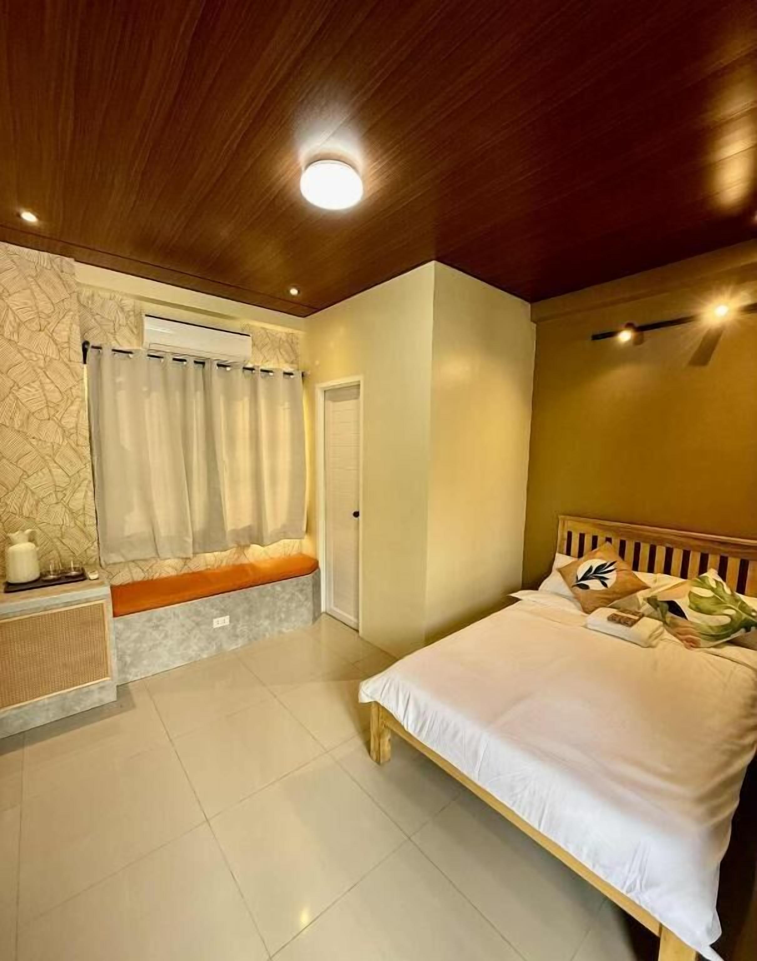 Un dormitorio con techo de madera, una cama con ropa blanca, un banco, una mesita de noche y un aire acondicionado de pared.