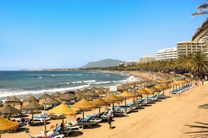 A beach with rows of sun loungers and thatched umbrellas, a clear blue sky, and a mountain in the distance. - Ipanema 610 - La Cala De Mijas, Andalucía (La Cala De Mijas)