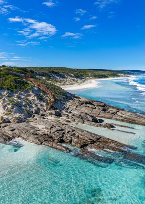 A coastal landscape with clear turquoise waters, rocky terrain, and a wooden staircase leading down to the beach.