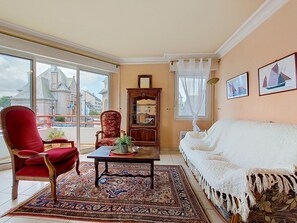 A room with a red chair, white sofa, wooden cabinet, and a large window with a view of buildings. - Hotel de Ville by Interhome (Perros Guirec)