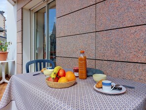 Ein Tisch mit Früchten, einer Flasche und einem Croissant auf einer gemusterten Tischdecke vor einem Gebäude mit einer Steinwand.