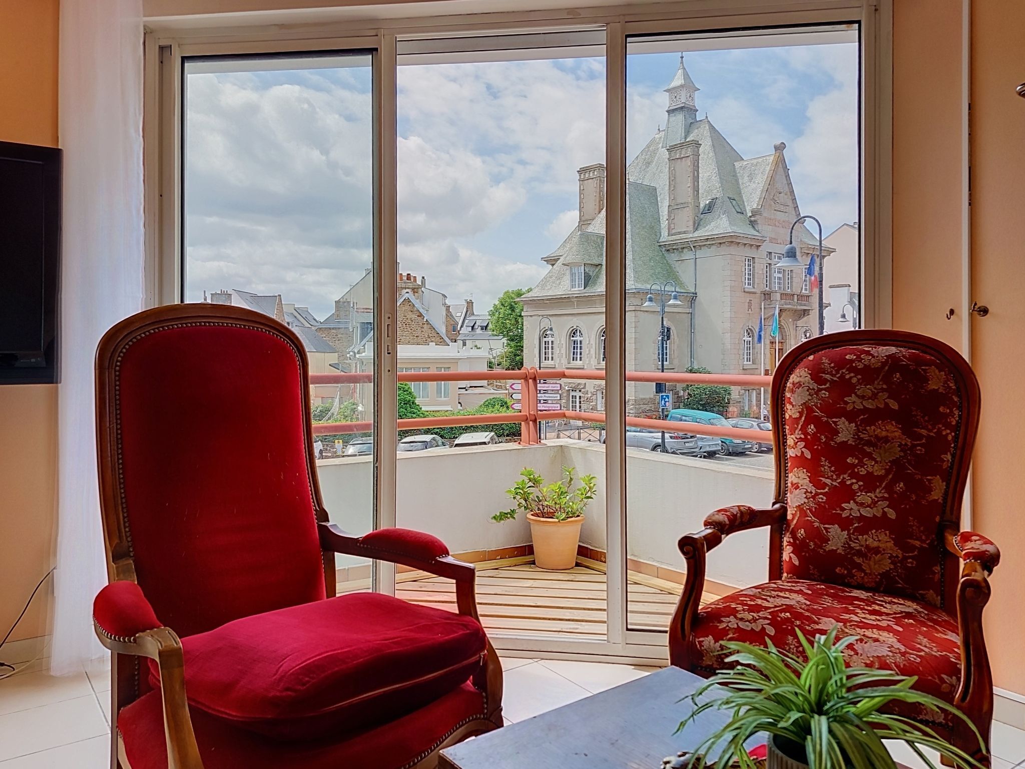 Ein Zimmer mit zwei roten Polstersesseln, einem kleinen Tisch und Blick auf ein historisches Gebäude durch ein großes Fenster.